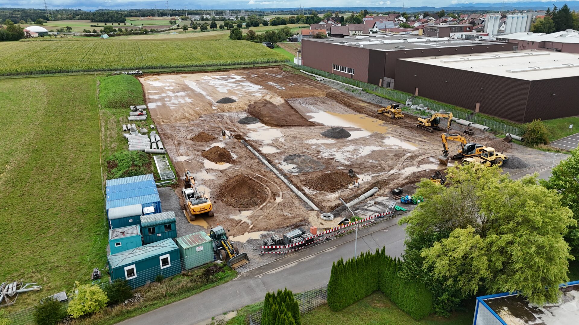 Luftaufnahme einer Baustelle mit Baggern, Erdarbeiten und Baumaschinen neben Industriegebäuden in ländlicher Gegend.